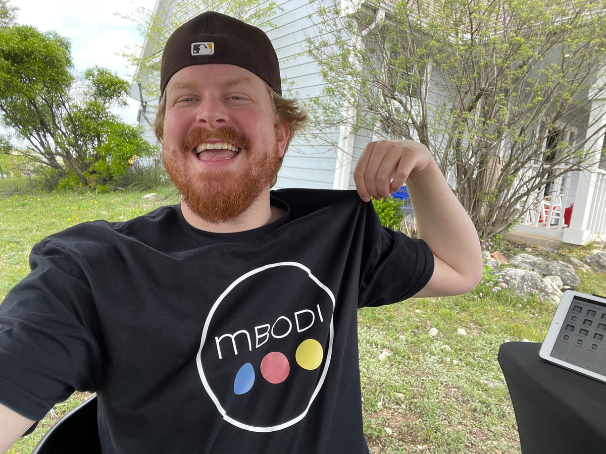 Mbodi creator wearing a black t-shirt with Mbodi logo, sitting outdoors at a farmers market.